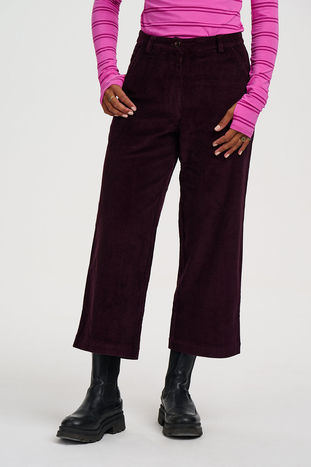 A model wearing dark plum corduroy cropped wide-leg pants, paired with a bright pink striped long-sleeve top and black ankle boots, against a plain light gray studio background.