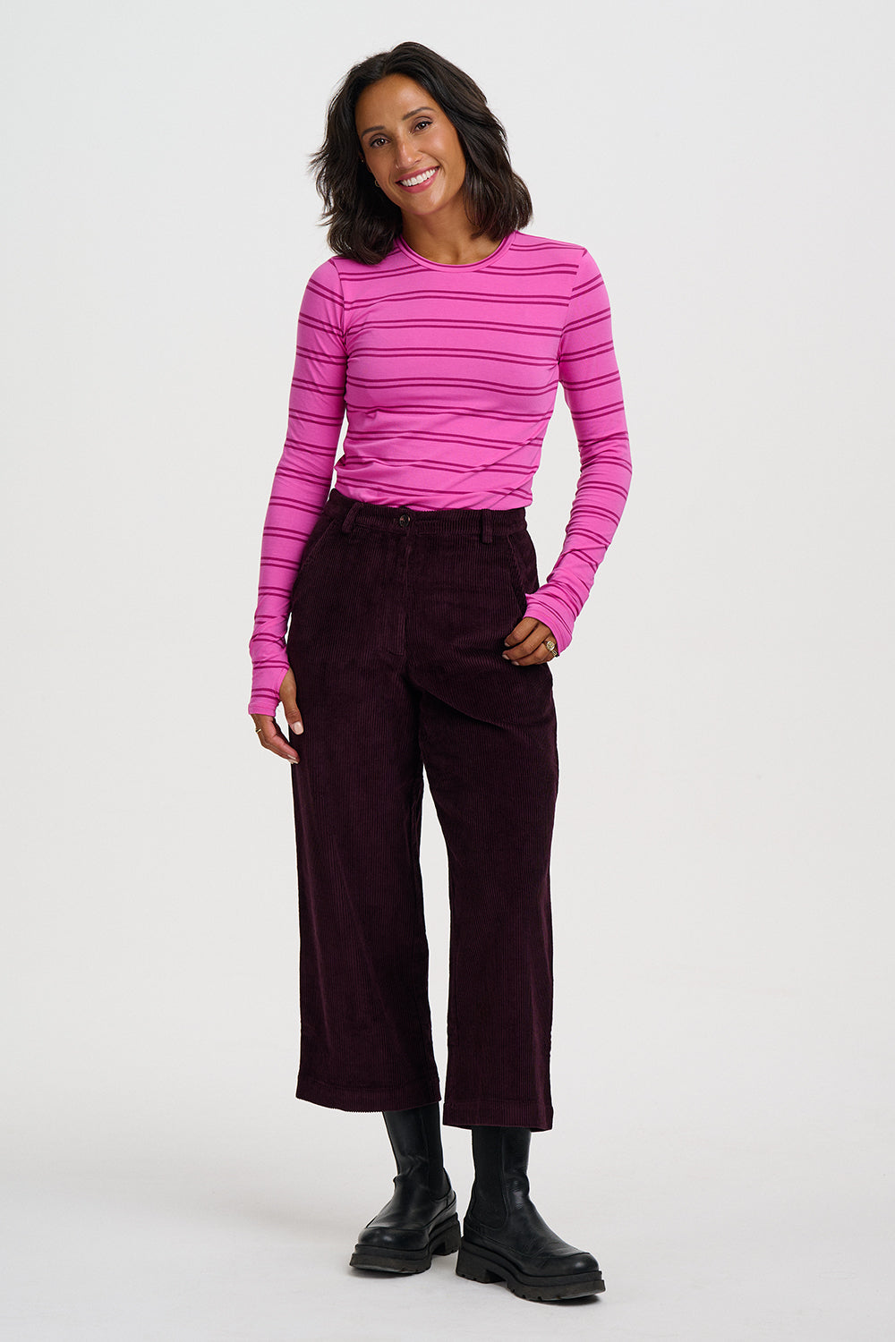 A woman modeling a bright pink and magenta striped long-sleeve top paired with dark burgundy cropped trousers and black boots, photographed against a light gray studio background.