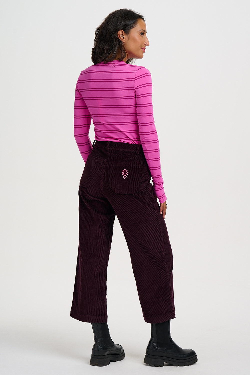 A woman shown from the back wearing a bright pink and magenta striped long-sleeve top, dark burgundy wide-leg corduroy pants with a small embroidered rose on the back pocket, and black boots, against a plain light gray studio background.