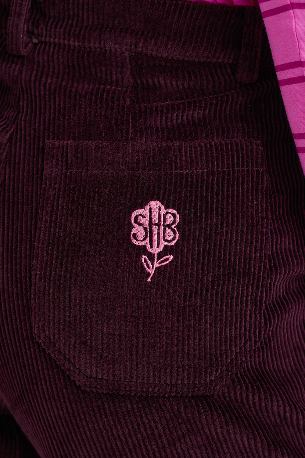 Close-up of dark plum corduroy trousers featuring a front pocket with a pink embroidered monogram logo.