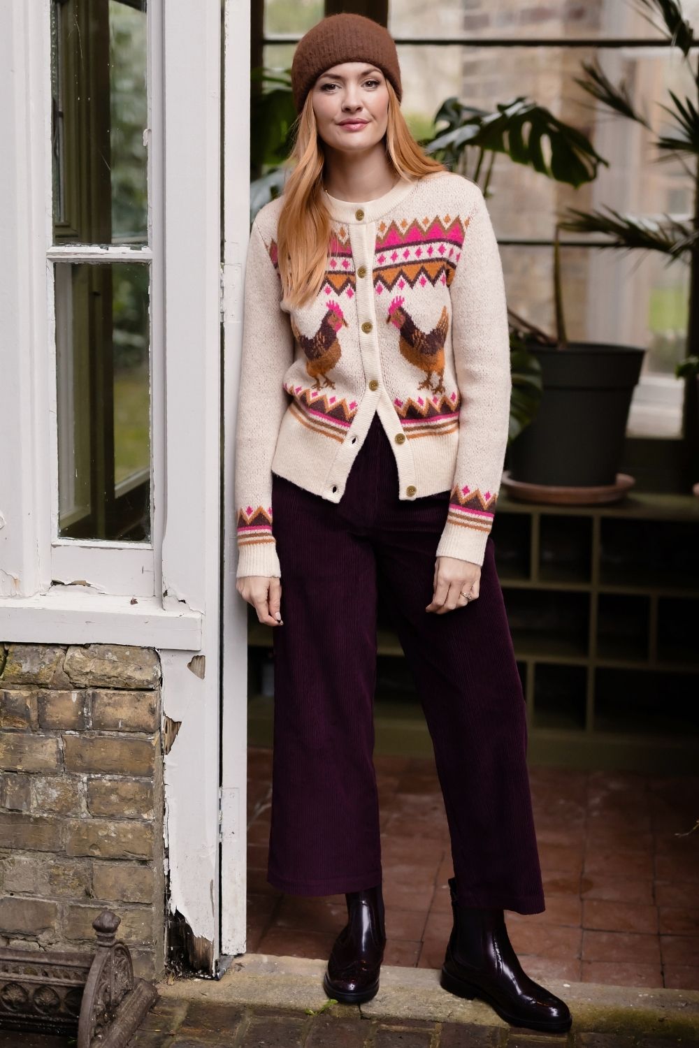A woman stands in a greenhouse doorway wearing a cream cardigan with a colorful Fair Isle pattern and rooster motifs, dark purple wide-leg trousers, black ankle boots, and a brown beanie.