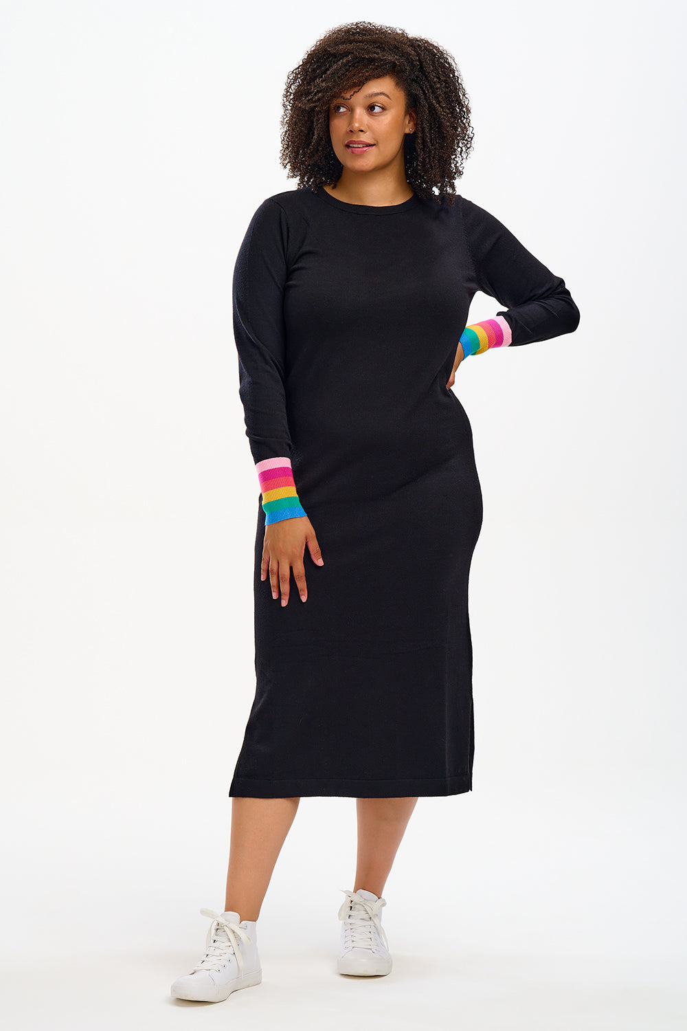 A model wearing a black knit dress with rainbow stripe cuffs on the sleeves, standing against a white background.
