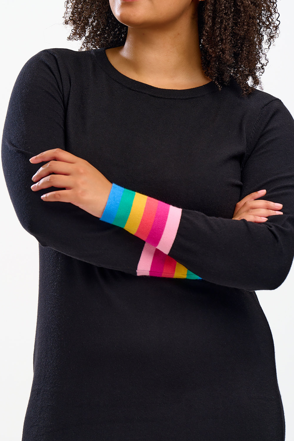 A model wearing a black knit dress with rainbow stripe cuffs on the sleeves, standing against a white background.