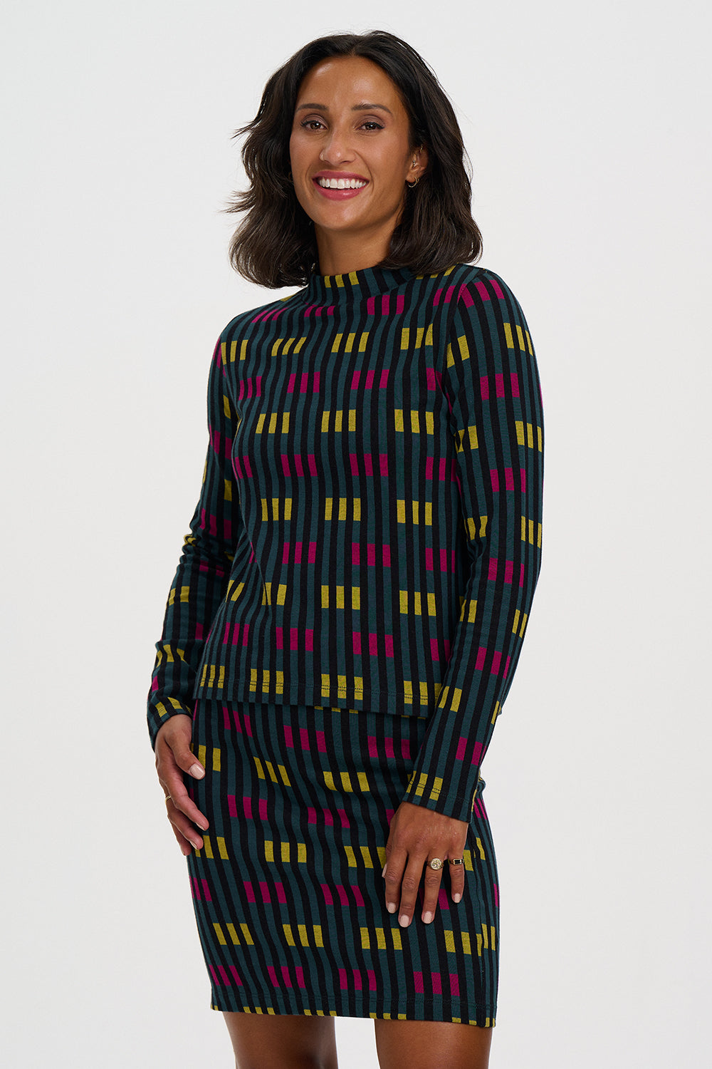 A woman modeling a long-sleeve mock-neck dress with a dark green base and a colorful geometric pattern of yellow and magenta vertical bars, set against a plain light gray background.