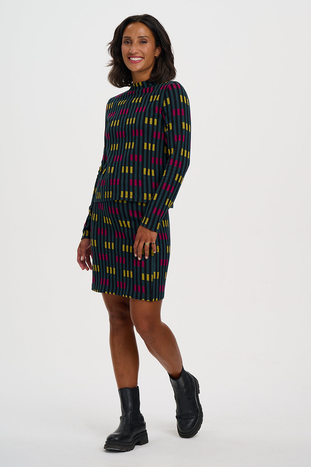 A woman modeling a long-sleeve ribbed knit mini dress with a vertical stripe pattern in black, magenta, and yellow, paired with black ankle boots, against a plain light gray studio background.