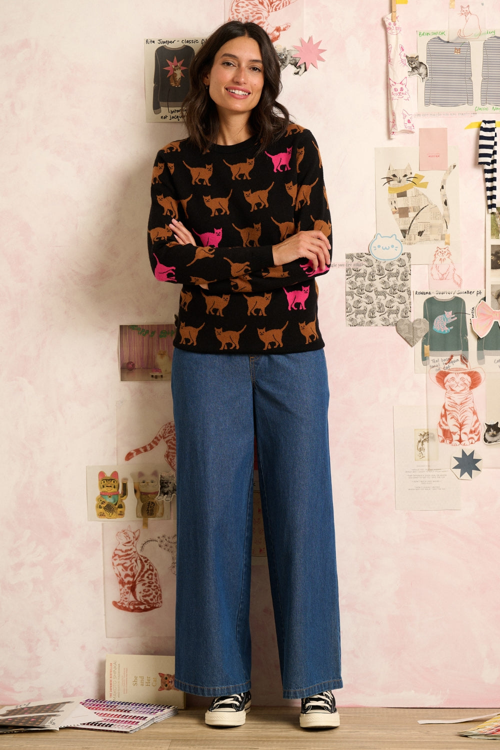 Woman wearing a black sweater with dog patterns and blue jeans standing in front of a wall with various items.