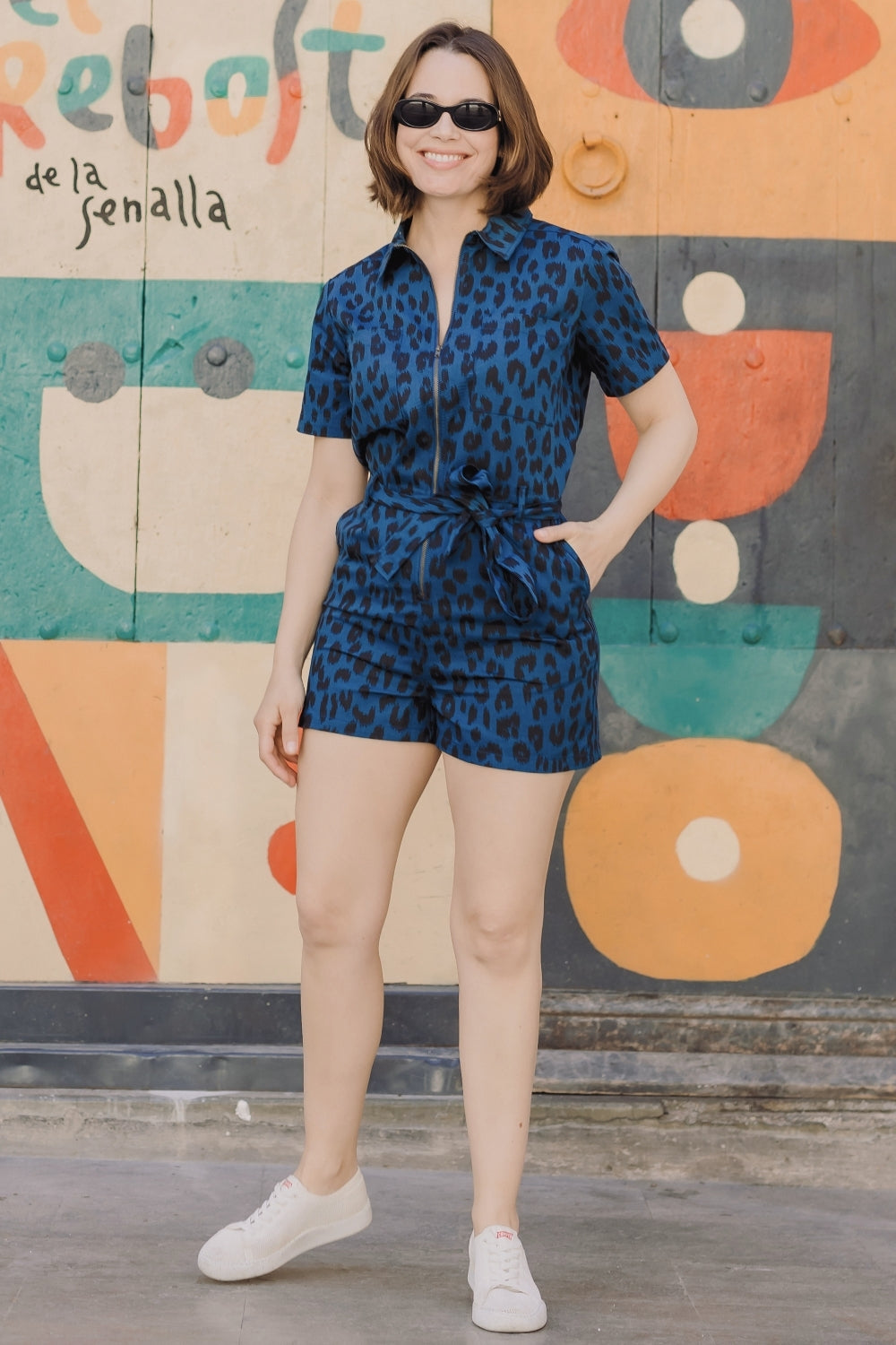 Woman in a blue patterned romper standing in front of a colorful mural.