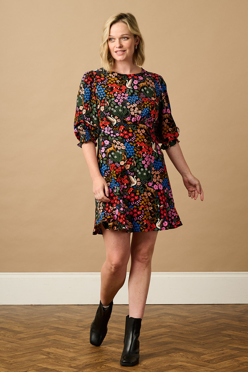 A woman standing and smiling, wearing a multicolored floral dress with half-length blouson sleeves and a frill hem skirt.