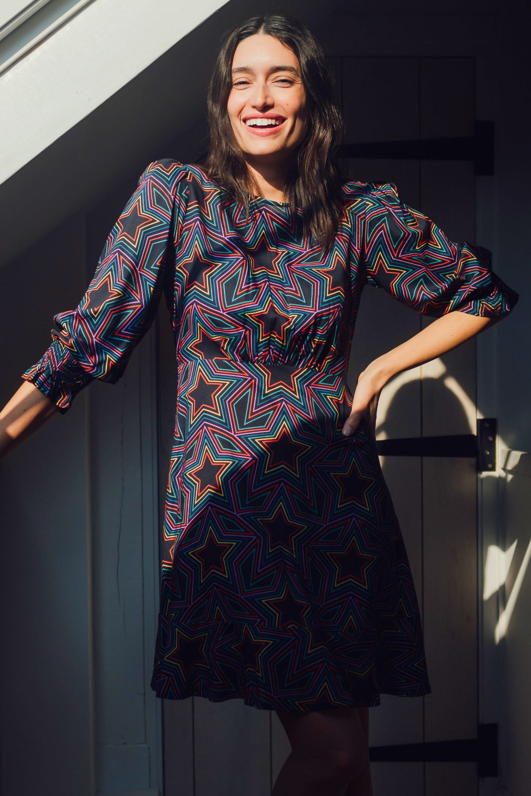 A woman standing in a doorway wearing a knee-length black dress with a multicolored star print and long blouson sleeves.