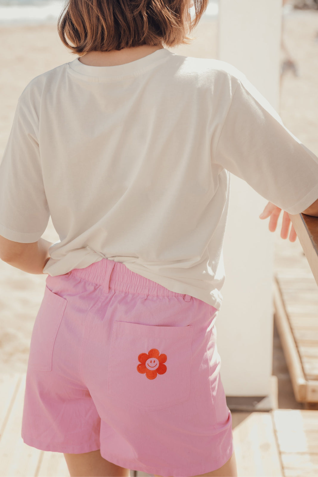 A pair of pink shorts with a red smiley flower pocket on the back, worn by a person viewed from the back.