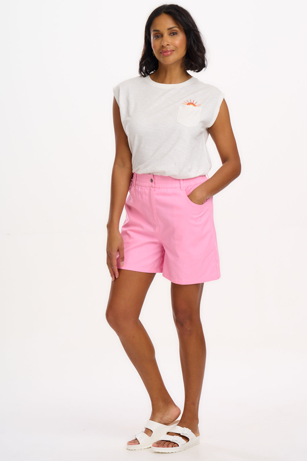 A woman standing, wearing bubblegum pink shorts with a smiley flower pocket and a white top.