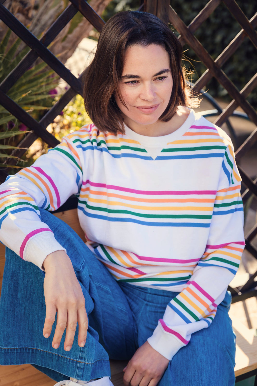 A woman sitting and wearing a long-sleeved sweatshirt with off-white and multicolored horizontal stripes and blue jeans.