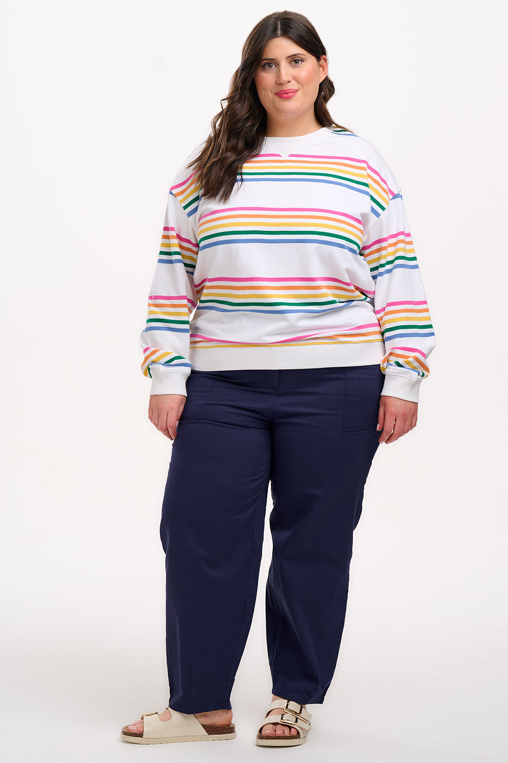A female model wearing an off-white sweatshirt with multicolored horizontal stripes in pink, orange, yellow, green, and blue, paired with blue jeans.