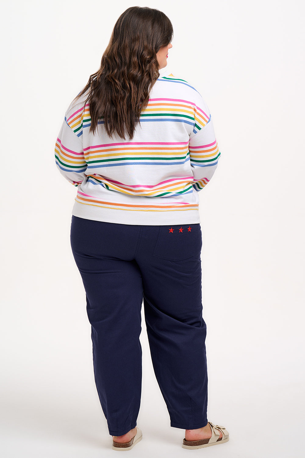 A female model wearing an off-white sweatshirt with multicolored horizontal stripes in pink, orange, yellow, green, and blue, paired with blue jeans.