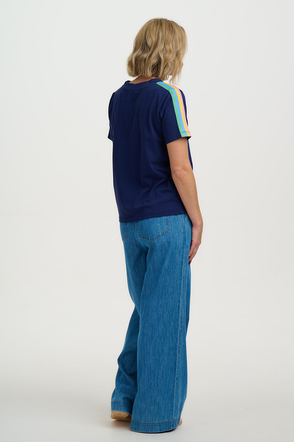 Person wearing a navy blue t-shirt with colorful stripes on sleeves and blue jeans against a white background