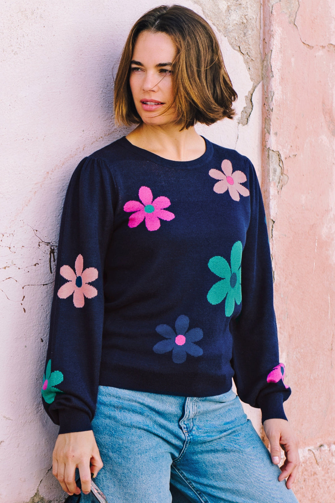 A woman wearing a navy blue jumper with a floral pattern consisting of large flowers in pink and green, paired with jeans.