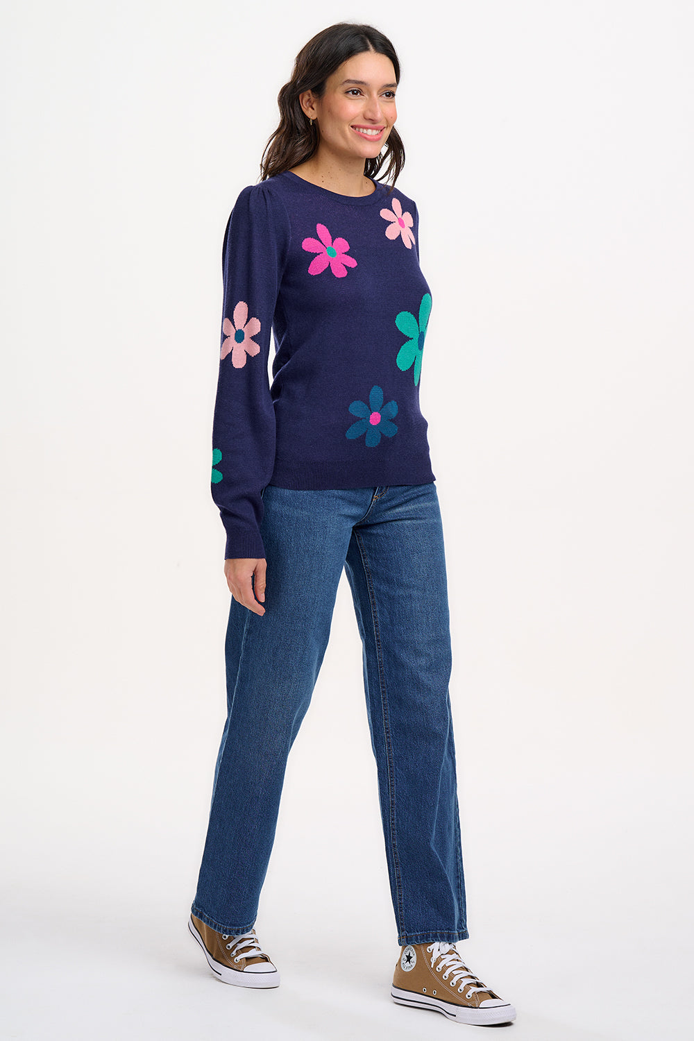 A woman wearing a navy blue jumper with a floral pattern consisting of large flowers in pink and green, paired with jeans.