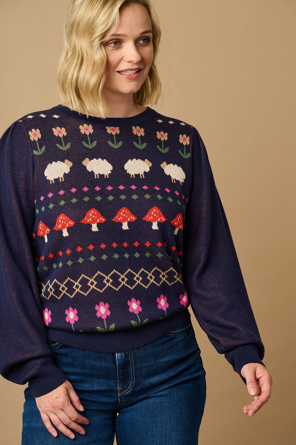 A woman wearing a navy blue jumper with a colorful fairisle pattern featuring sheep, toadstools, and flowers, paired with black jeans.