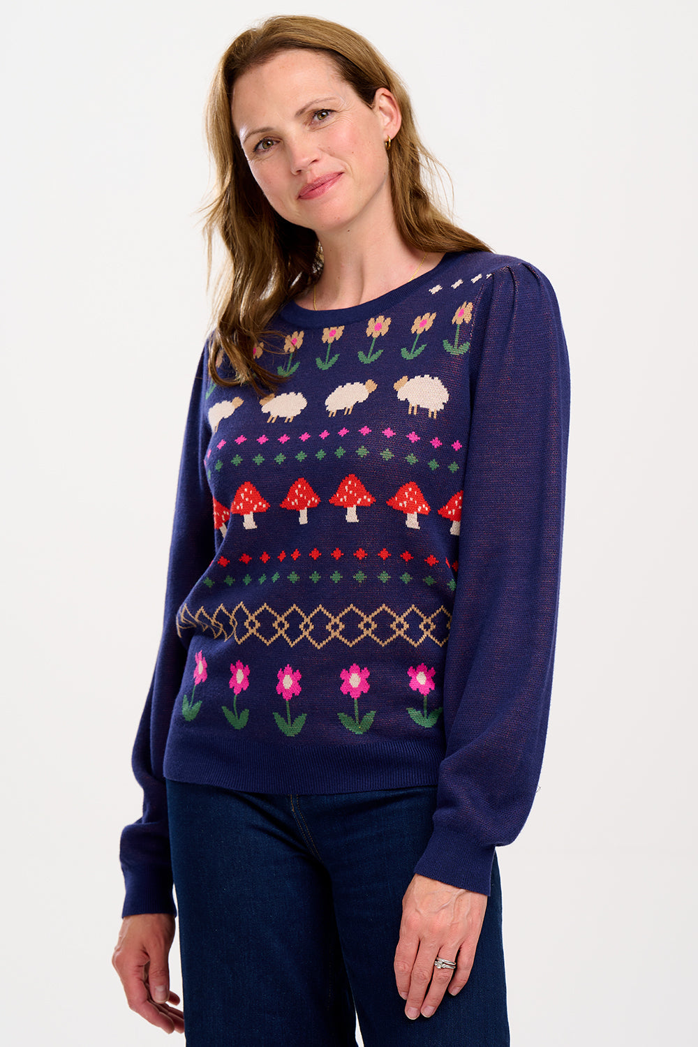 A woman wearing a navy blue jumper with a colorful fairisle pattern featuring sheep, toadstools, and flowers, paired with black jeans.
