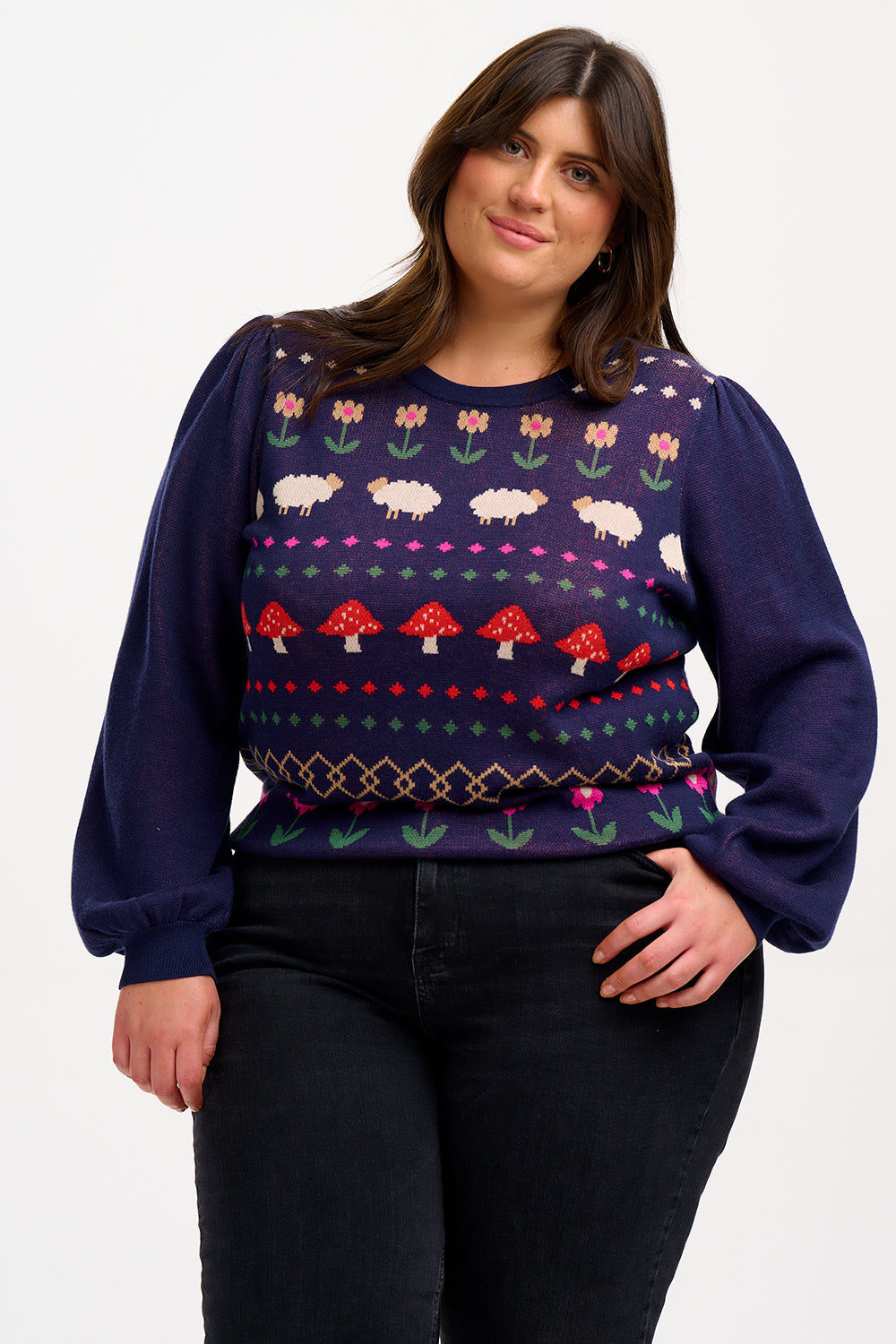 A woman wearing a navy blue jumper with a colorful fairisle pattern featuring sheep, toadstools, and flowers, paired with black jeans.