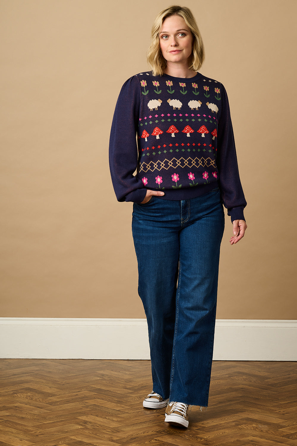 A woman wearing a navy blue jumper with a colorful fairisle pattern featuring sheep, toadstools, and flowers, paired with black jeans.