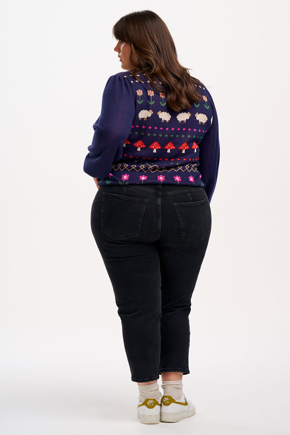 A woman wearing a navy blue jumper with a colorful fairisle pattern featuring sheep, toadstools, and flowers, paired with black jeans.