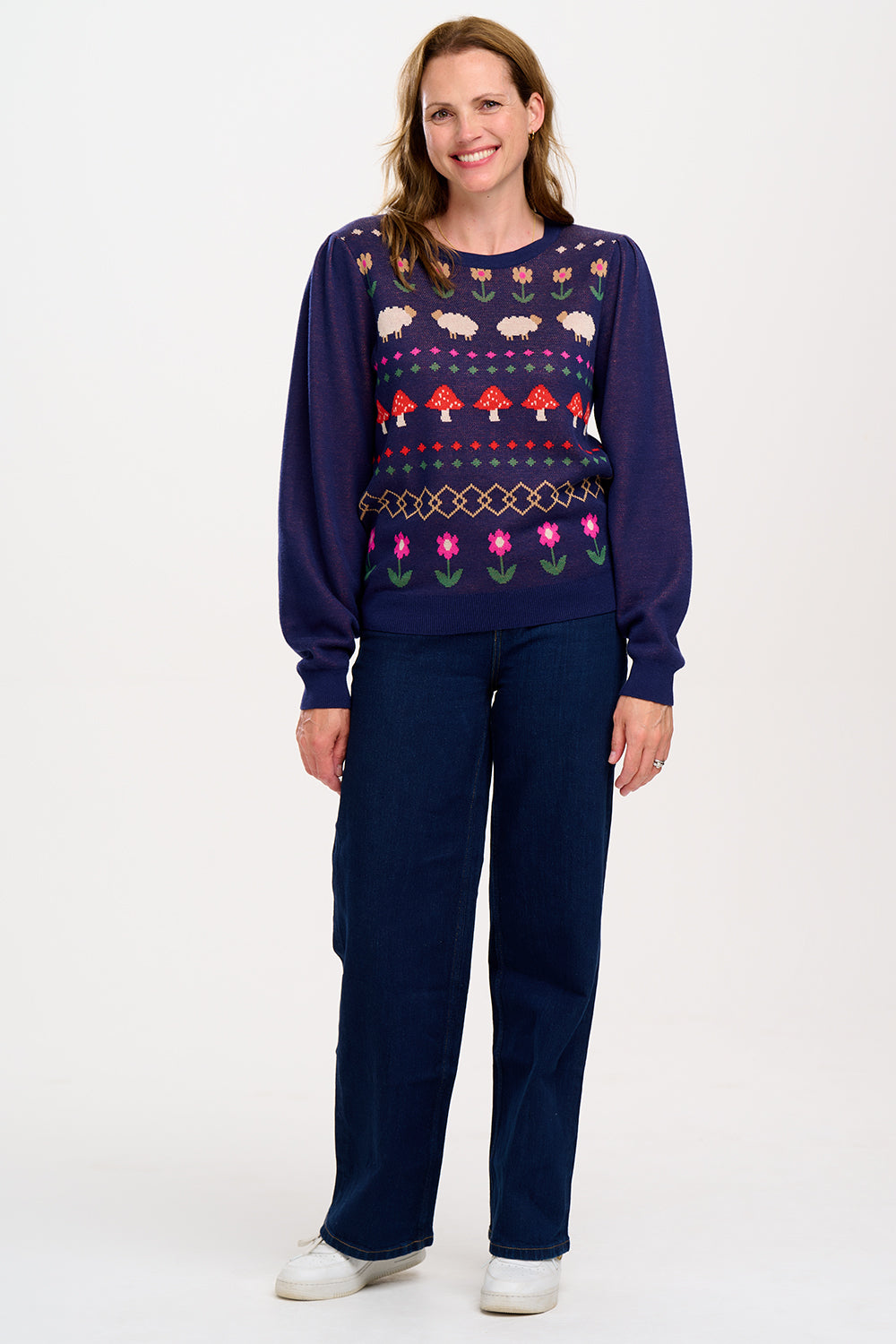 A woman wearing a navy blue jumper with a colorful fairisle pattern featuring sheep, toadstools, and flowers, paired with black jeans.