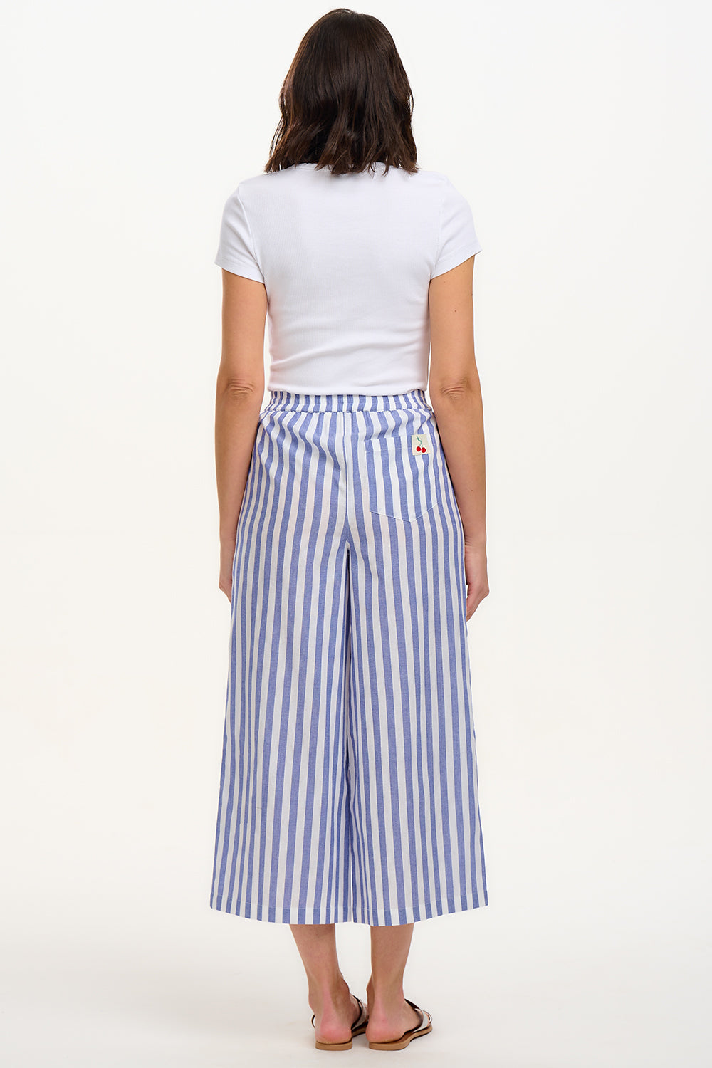 A woman modeling wide leg cropped trousers with an all-over bakers stripe in white and blue, paired with a white t-shirt and white sandals.