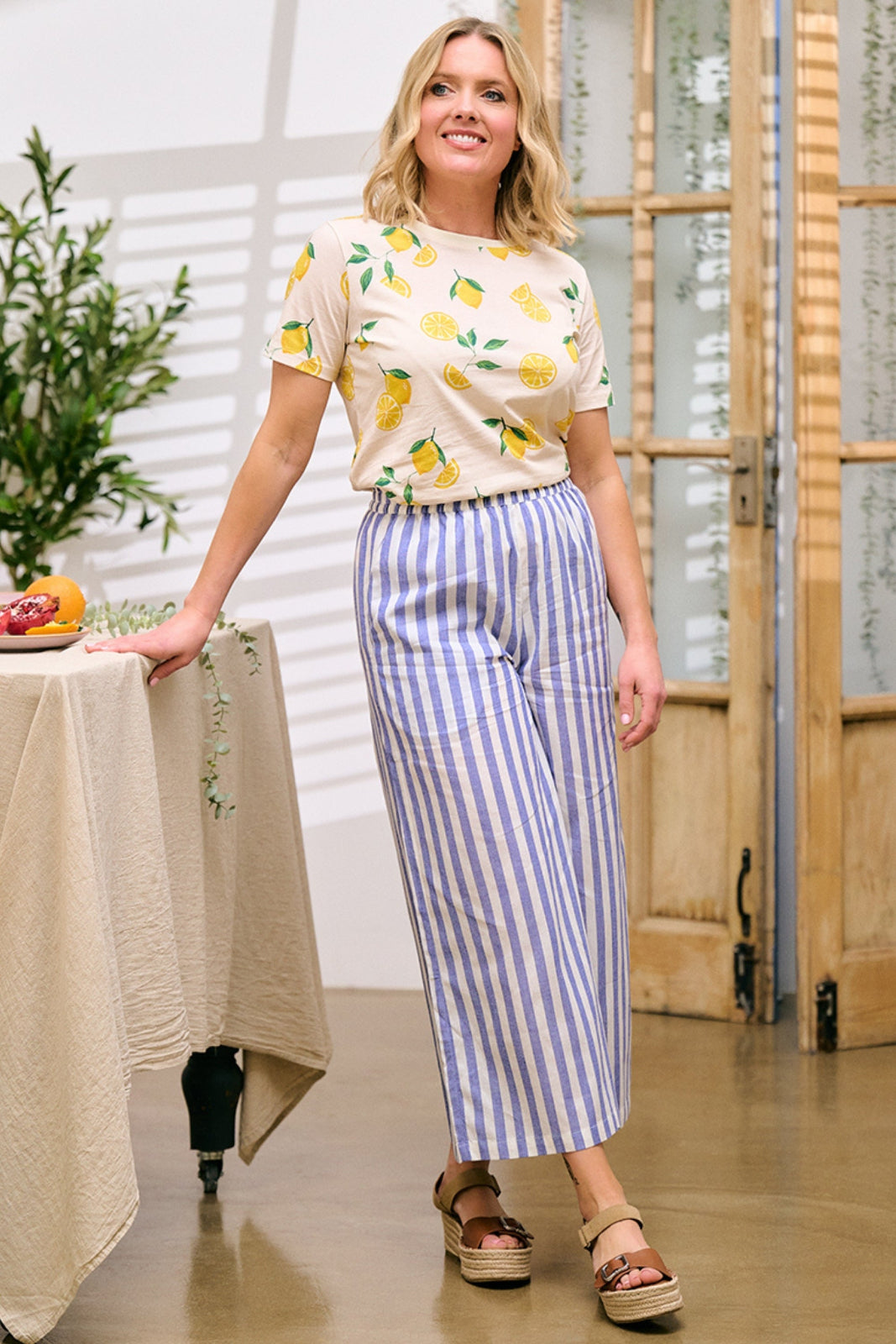 A woman standing in a room wearing wide leg cropped trousers with a bakers stripe in off-white and blue, paired with a yellow and white patterned top.