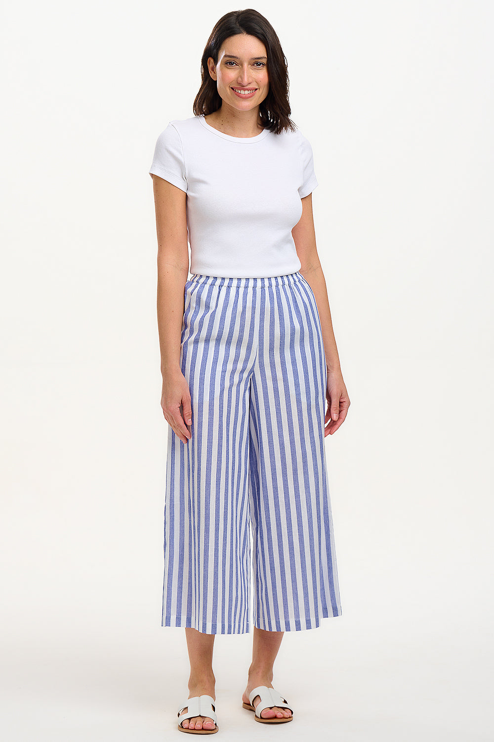 A woman modeling wide leg cropped trousers with an all-over bakers stripe in white and blue, paired with a white t-shirt and white sandals.