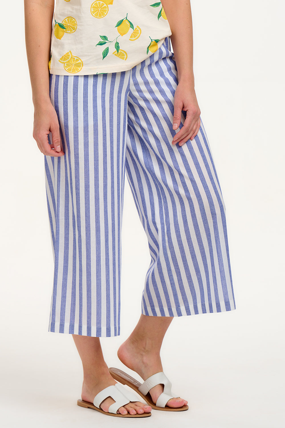 A woman modeling wide leg cropped trousers with an all-over bakers stripe in white and blue, paired with a white t-shirt and white sandals.