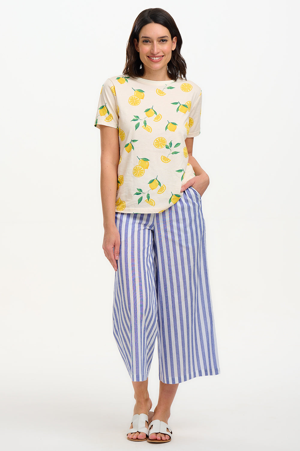 A woman modeling wide leg cropped trousers with an all-over bakers stripe in white and blue, paired with a white t-shirt and white sandals.