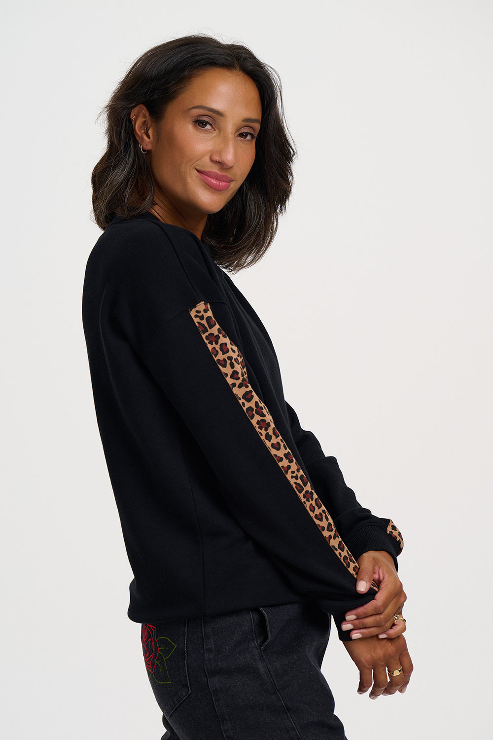 A woman wearing a black crewneck sweatshirt with a brown and black leopard print stripe detail down the right sleeve, paired with dark jeans, photographed against a solid light gray background.