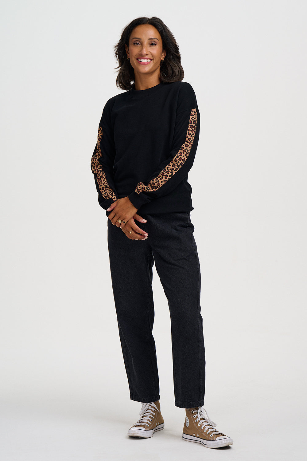 A woman modeling a black long-sleeve sweatshirt with leopard-print patterned sleeves, paired with black pants and brown sneakers, against a plain light gray studio background.