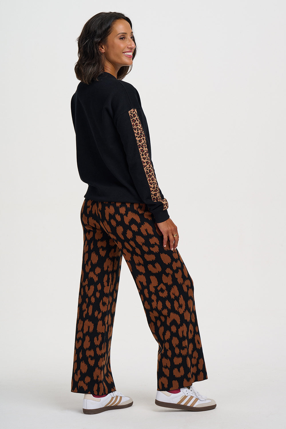 A woman modeling a black long-sleeve top with a leopard-print sleeve stripe and matching wide-leg leopard-print pants, paired with white sneakers, against a plain light gray studio background.