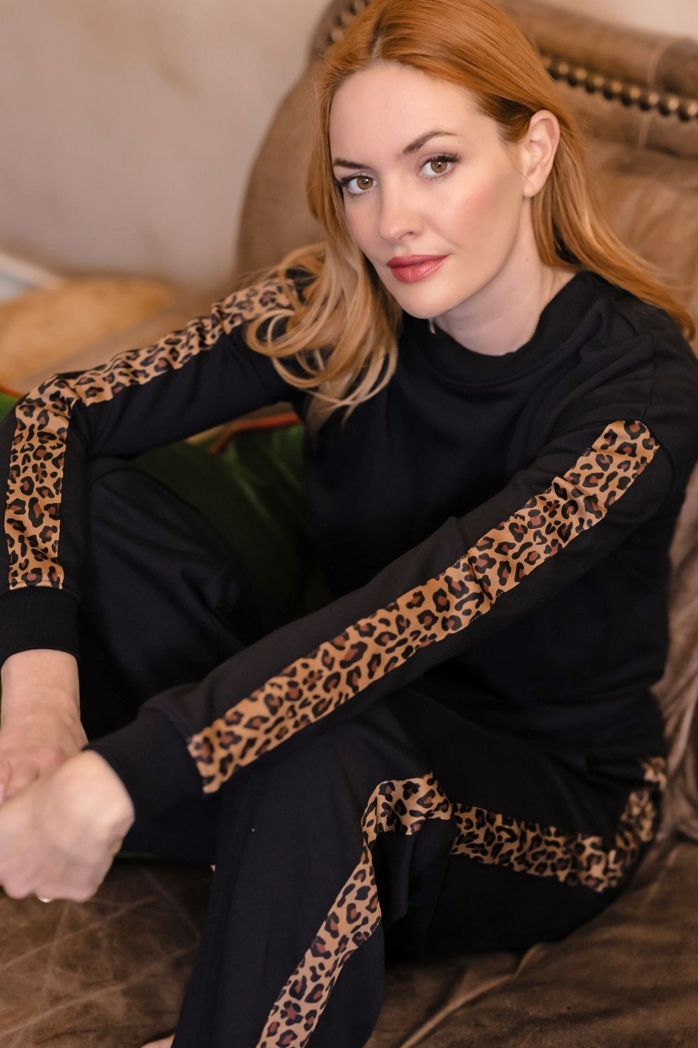 A woman with red hair sits on a brown leather couch wearing a black long-sleeve top and matching pants with leopard print stripes down the sleeves and along the hem.