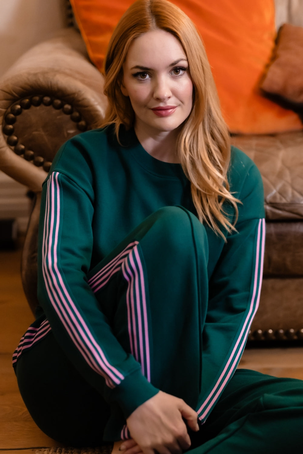 A woman with long red hair sits cross-legged on a wooden floor in a cozy living room setting, wearing a dark green sweatshirt and matching sweatpants with pink and white stripes down the sleeves and legs.