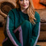 A woman with long red hair sits cross-legged on a wooden floor in a cozy living room setting, wearing a dark green sweatshirt and matching sweatpants with pink and white stripes down the sleeves and legs.
