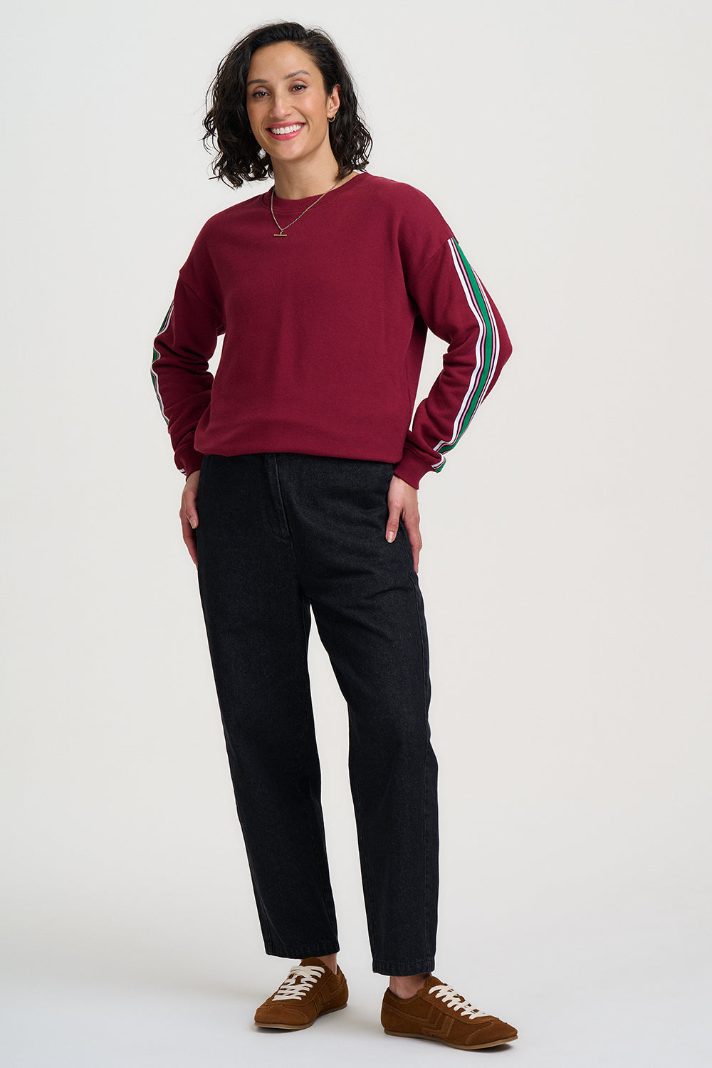 Person wearing a maroon sweatshirt with colorful stripes on sleeves, black pants, and brown shoes against a white background