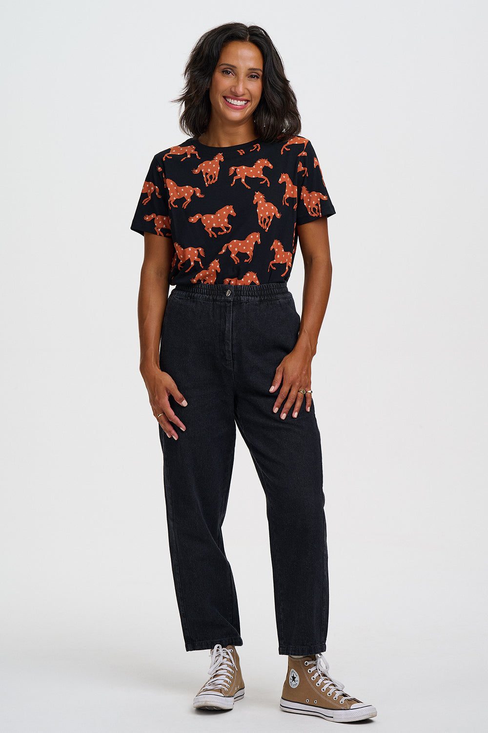 A woman modeling a black short-sleeve t-shirt with an orange horse pattern, paired with black jeans and brown Converse sneakers, against a light gray studio background.