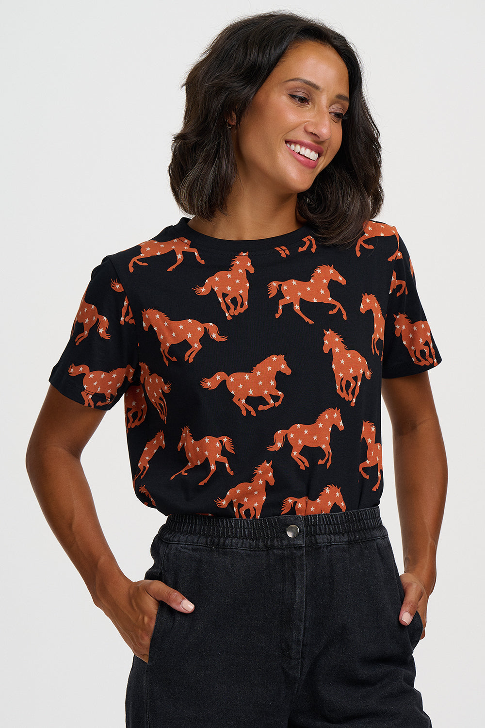 A woman wearing a black short-sleeve t-shirt with an all-over print of brown horses with white spots, paired with dark denim jeans, against a light gray studio background.