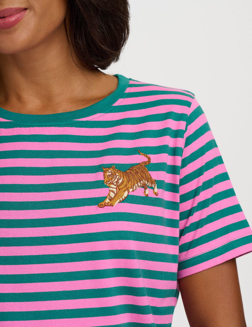 A close-up of a model wearing a pink and teal striped short-sleeve t-shirt with an embroidered tiger on the chest.