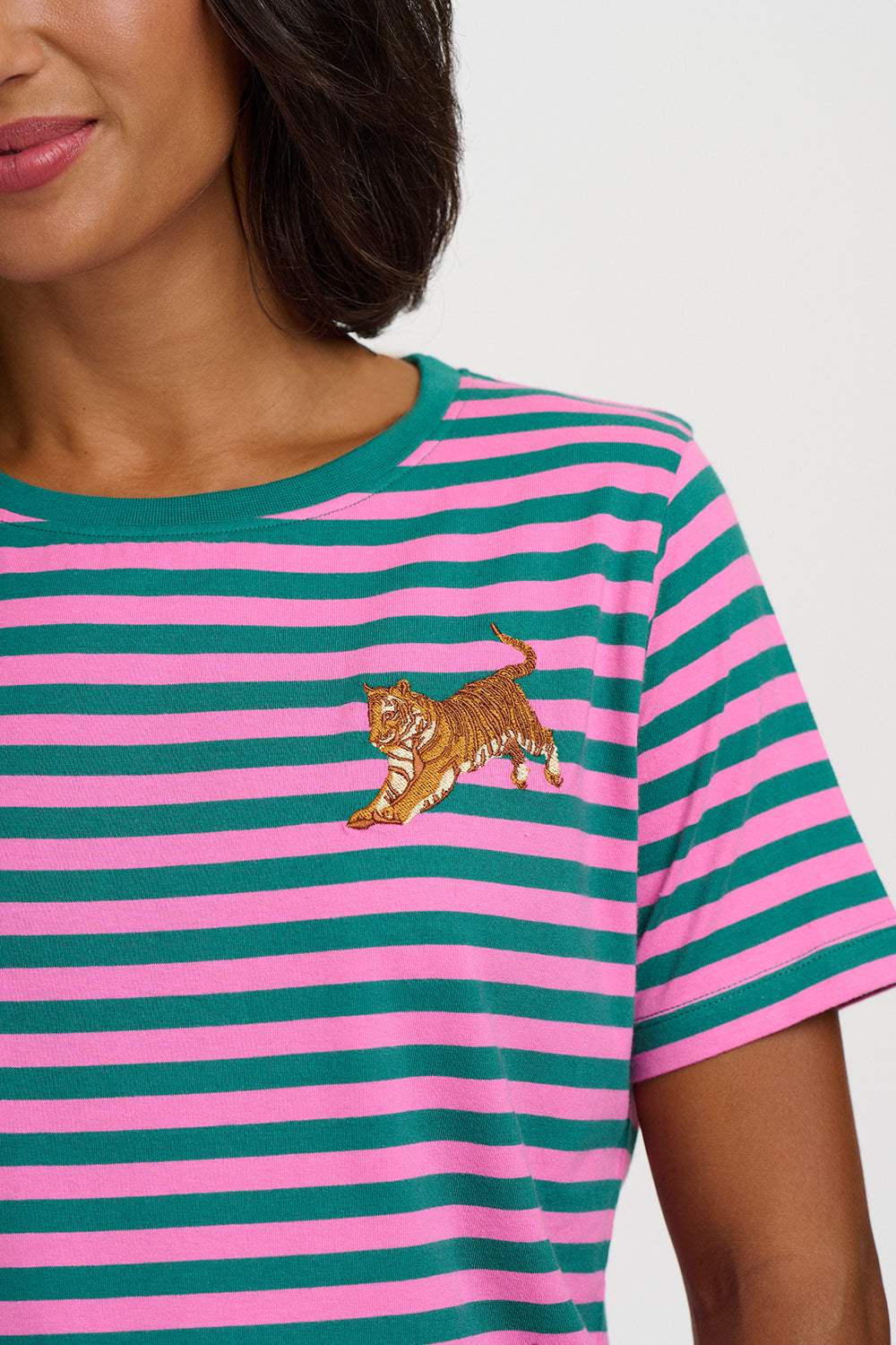 A close-up of a model wearing a pink and teal striped short-sleeve t-shirt with an embroidered tiger on the chest.