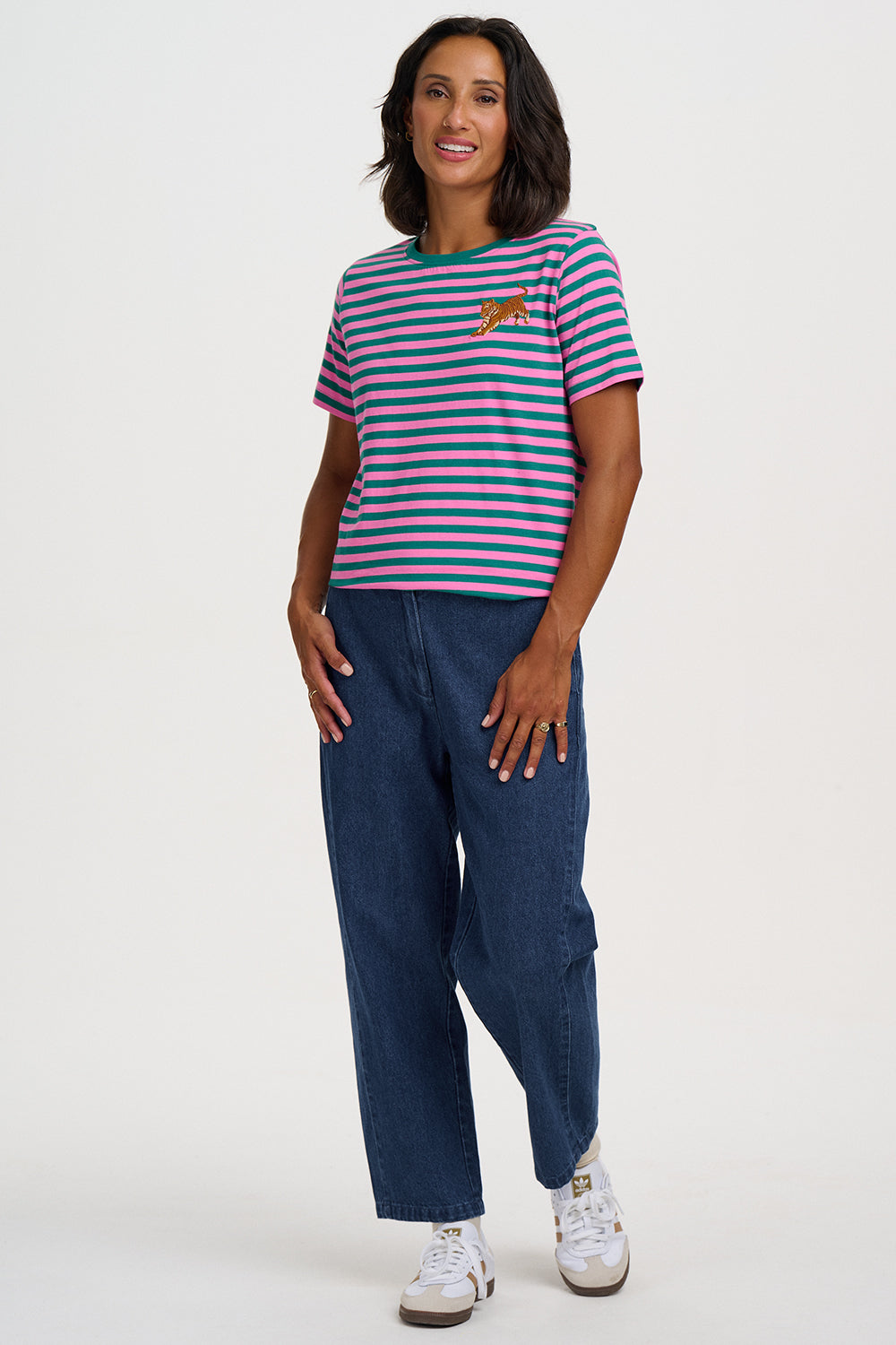 A woman modeling a short-sleeve crewneck t-shirt with horizontal navy, pink, and white stripes, featuring a small embroidered tiger on the left chest. She is wearing dark blue wide-leg cargo pants and white sneakers against a plain light gray background.