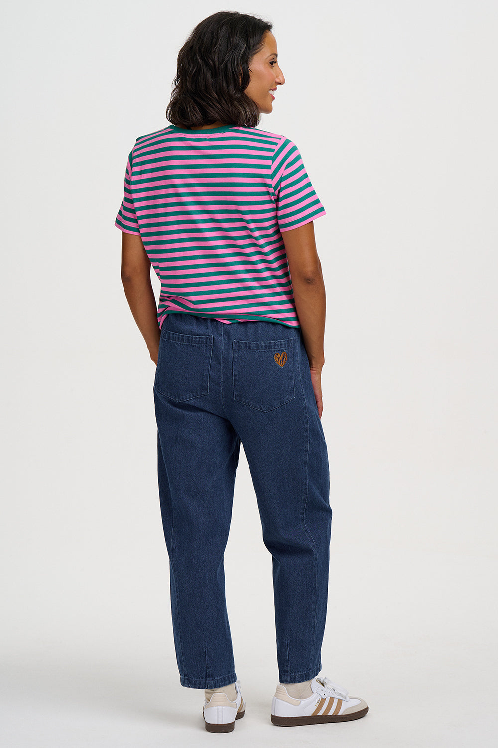 A woman shown from the back wearing a short-sleeve striped t-shirt with blue, pink, and green stripes, dark blue jeans, and white sneakers, against a plain light gray background.