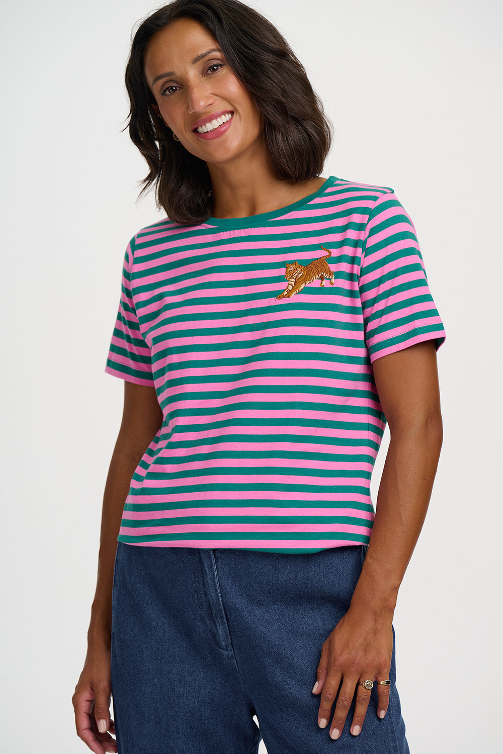 A woman wearing a short-sleeve striped t-shirt with pink and teal horizontal stripes, featuring a small embroidered leopard on the left chest. She is also wearing blue jeans against a plain light gray studio background.