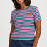 A woman wearing a short-sleeve striped t-shirt with pink and teal horizontal stripes, featuring a small embroidered leopard on the left chest. She is also wearing blue jeans against a plain light gray studio background.