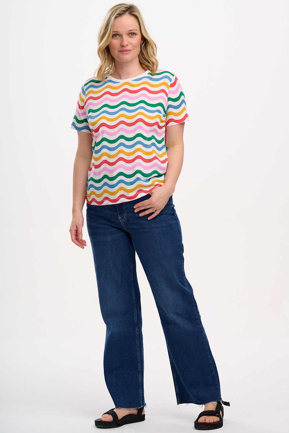 A woman smiling and wearing a multicolored wavy rainbow stripe t-shirt with a crew neck.