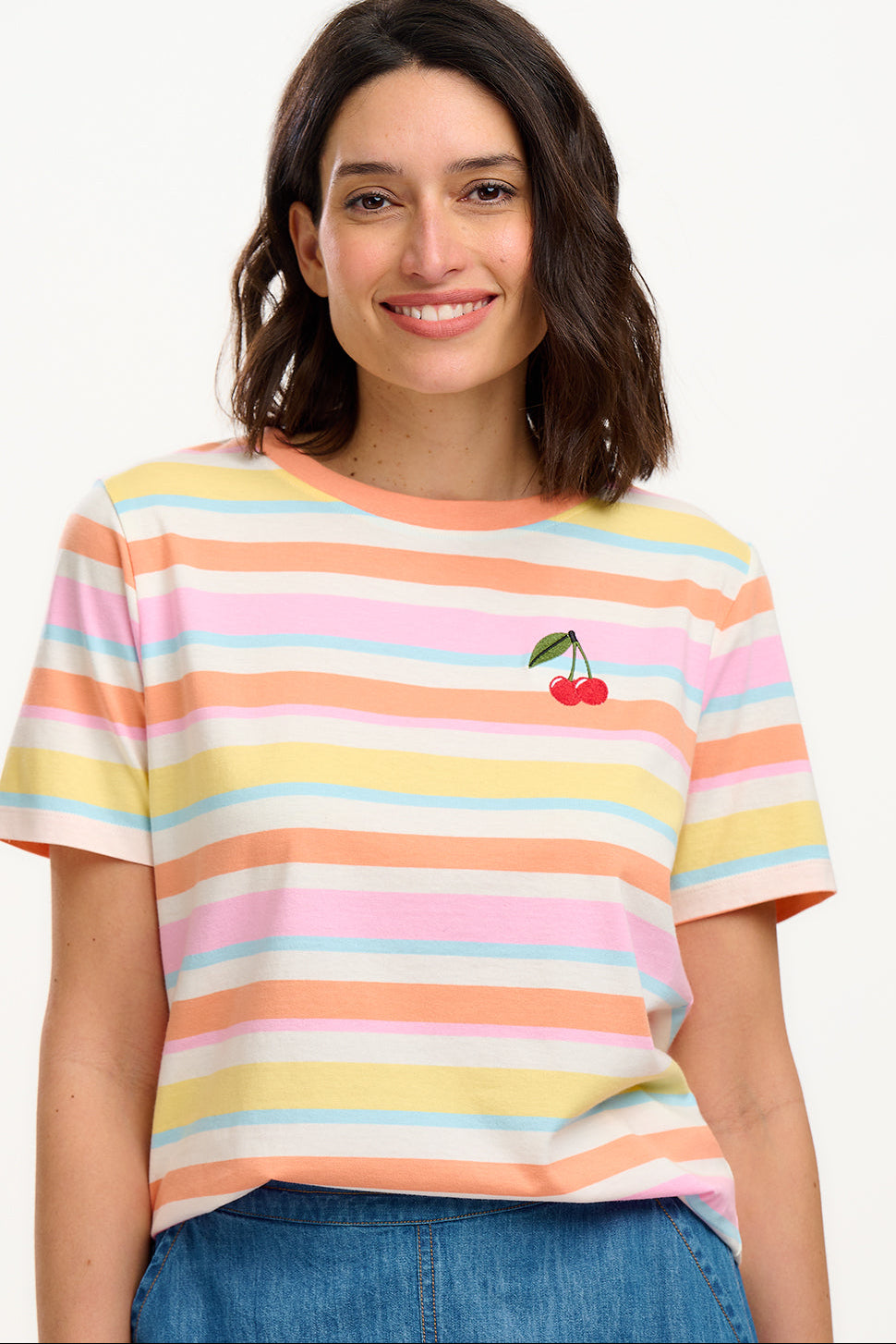 A woman modeling a multicolored striped t-shirt with a small embroidered cherry on the chest, paired with a blue denim skirt.