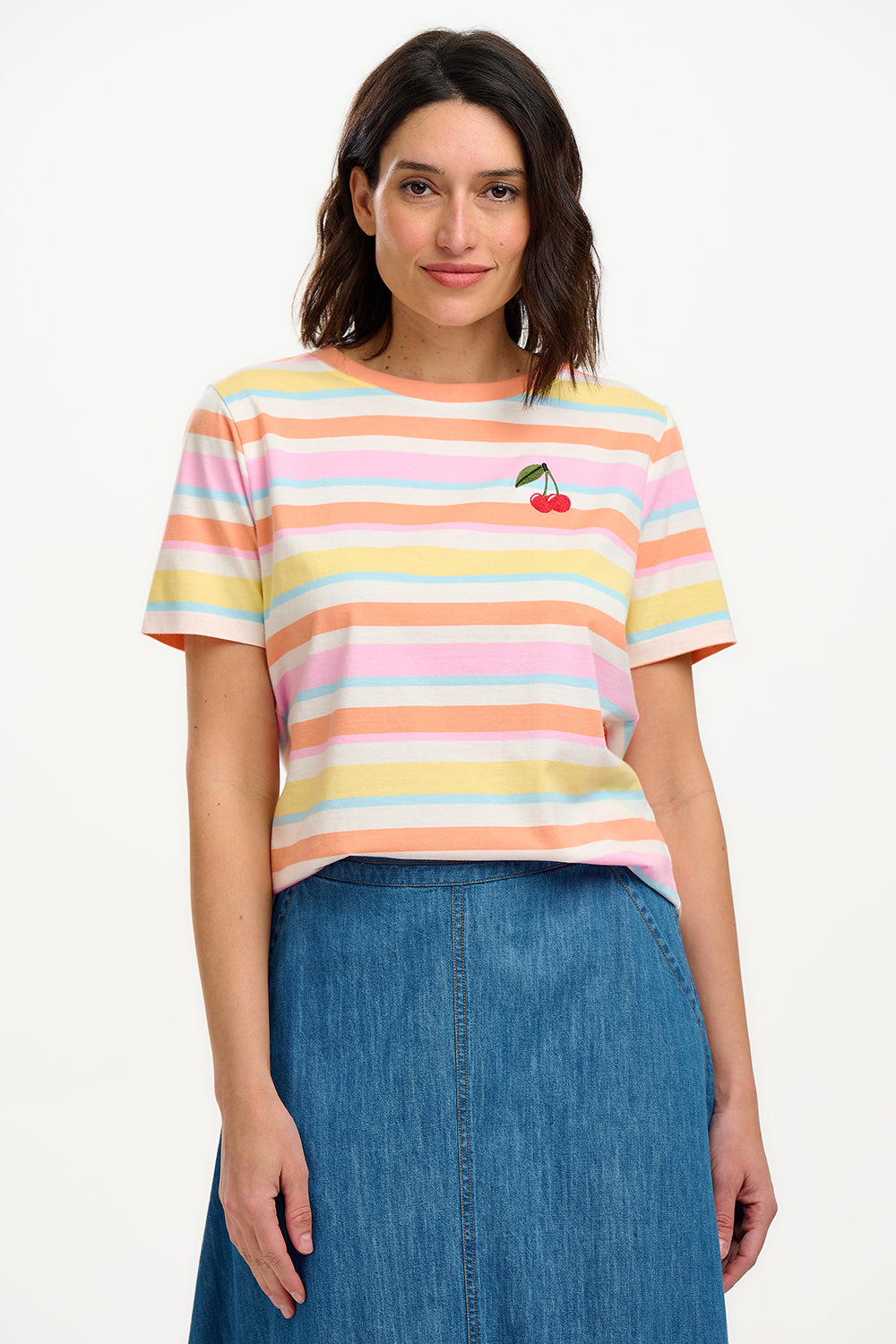 A woman modeling a multicolored striped t-shirt with a small embroidered cherry on the chest, paired with a blue denim skirt.